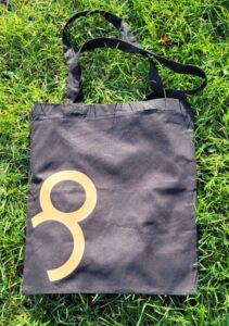 Auf dem Foto ist eine cremefarbene Stofftasche mit langen Henkeln zu sehen. Die Tasche liegt auf grünem Gras mit einigen gelben Blüten im Hintergrund. Auf der Vorderseite der Tasche befindet sich ein aufgedruckter Text sowie das Logo von der Bafep8..