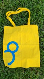 Auf dem Foto ist eine cremefarbene Stofftasche mit langen Henkeln zu sehen. Die Tasche liegt auf grünem Gras mit einigen gelben Blüten im Hintergrund. Auf der Vorderseite der Tasche befindet sich ein aufgedruckter Text sowie das Logo von der Bafep8..
